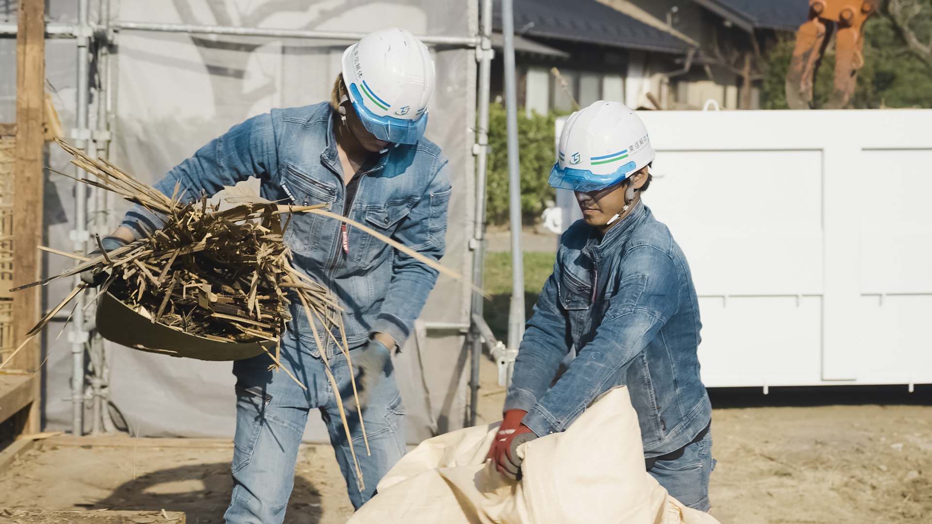 ヘルメットと作業着姿の2人の作業員が、協力して枯れ草などの廃材を大きな袋に片付けている様子